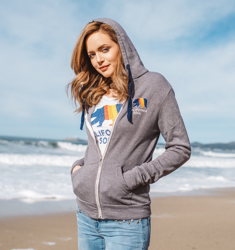 May include: A grey zip-up hoodie with a California bear logo and the text "California Soul". The model is wearing the hoodie with the zipper pulled up and is standing on a beach.