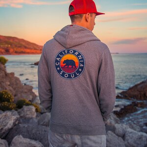 May include: A man wearing a grey hoodie with a California Soul logo featuring a bear and a sunset graphic. He is wearing a red mesh baseball cap and blue jeans. The man is standing on a rocky beach with the sea in the background.