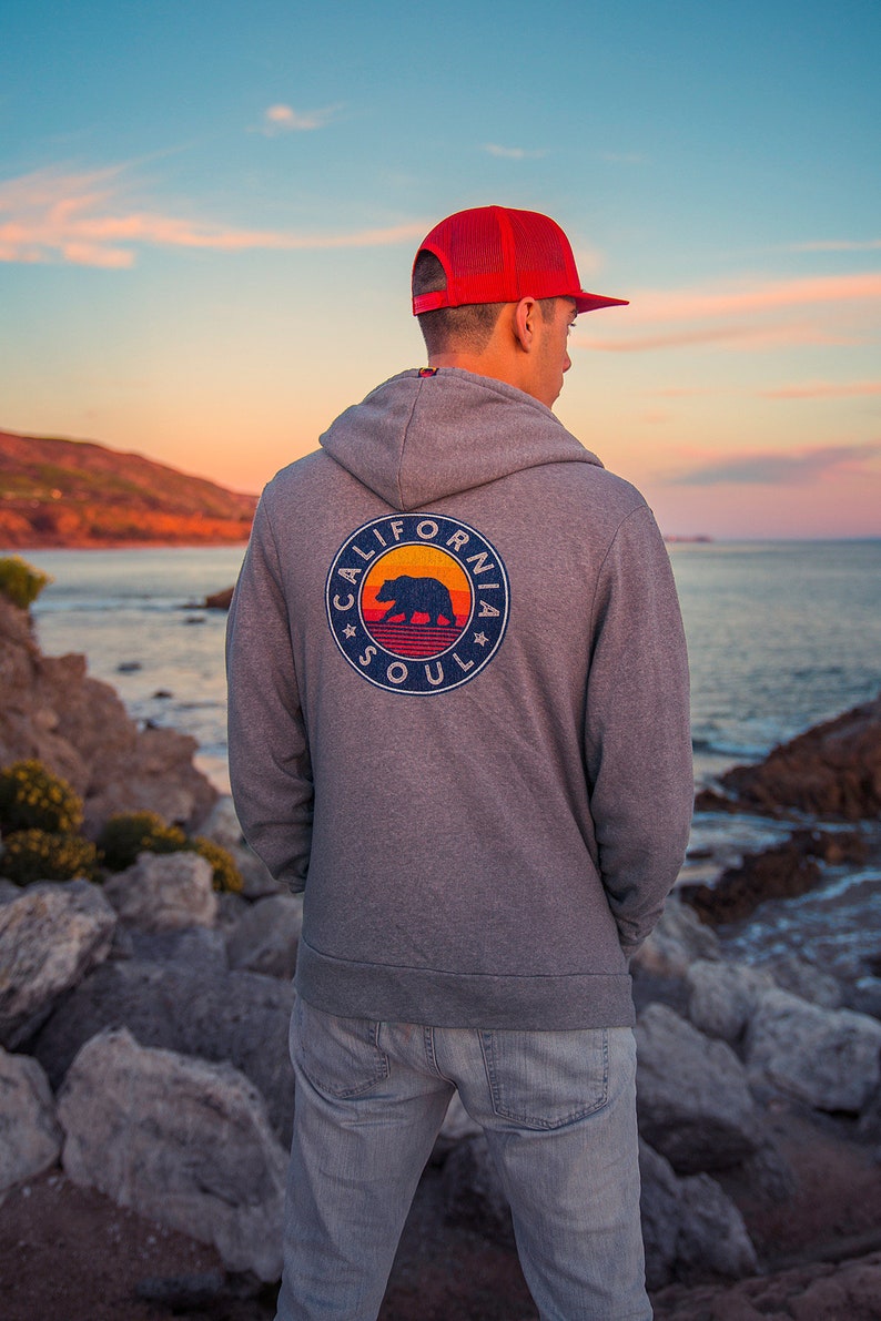 May include: A man wearing a grey hoodie with a California Soul logo featuring a bear and a sunset graphic. He is wearing a red mesh baseball cap and blue jeans. The man is standing on a rocky beach with the sea in the background.