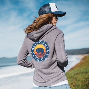May include: A grey hooded sweatshirt with a graphic of a bear and the text "California Soul" on the back. The sweatshirt has a white and blue baseball cap on top.