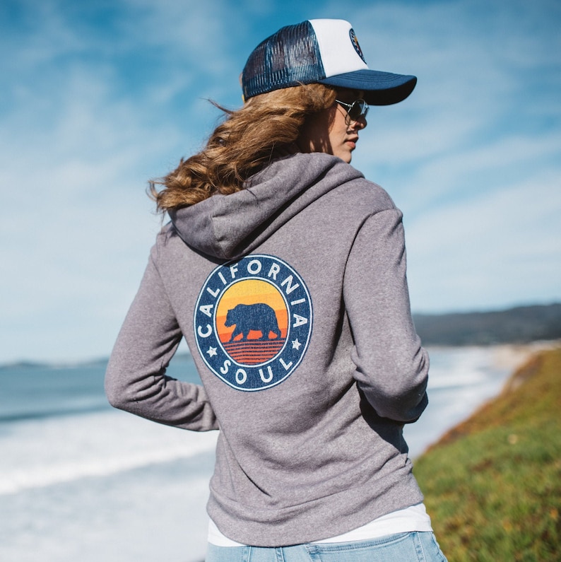 May include: A grey hooded sweatshirt with a graphic of a bear and the text "California Soul" on the back. The sweatshirt has a white and blue baseball cap on top.