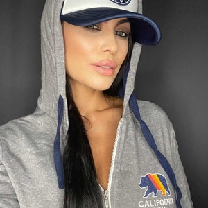 May include: A grey zip-up hoodie with a navy blue zipper and drawstrings. The hoodie features a California Soul logo with a bear and a sunset on the front. The model is wearing a white and navy blue trucker hat with a California Soul logo.