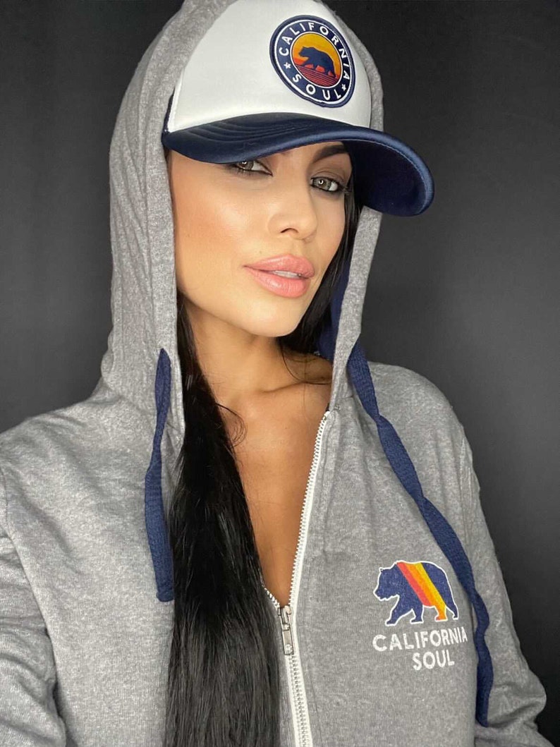 May include: A grey zip-up hoodie with a navy blue zipper and drawstrings. The hoodie features a California Soul logo with a bear and a sunset on the front. The model is wearing a white and navy blue trucker hat with a California Soul logo.