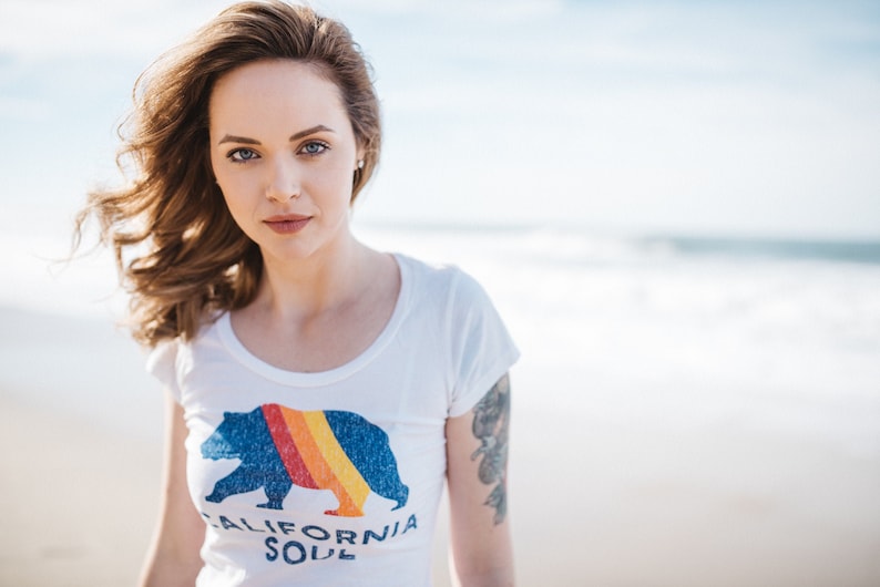May include: A woman with long brown hair is wearing a white t-shirt with a blue bear graphic and the text "CALIFORNIA SOUL".