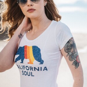 May include: A woman wearing a white t-shirt with a blue bear graphic and the text "CALIFORNIA SOUL". She is wearing sunglasses and has a tattoo on her arm.