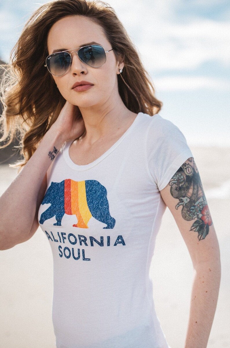 May include: A woman wearing a white t-shirt with a blue bear graphic and the text "CALIFORNIA SOUL". She is wearing sunglasses and has a tattoo on her arm.