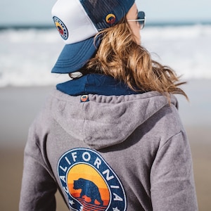 May include: A grey hooded sweatshirt with a blue and white California Soul logo featuring a bear and a sunset. The logo is on the back of the sweatshirt. The person is wearing a blue and white trucker hat.