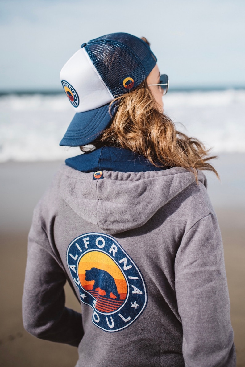 May include: A grey hooded sweatshirt with a blue and white California Soul logo featuring a bear and a sunset. The logo is on the back of the sweatshirt. The person is wearing a blue and white trucker hat.
