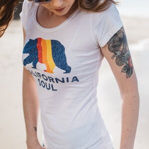 May include: A white t-shirt with a blue bear graphic and the text "CALIFORNIA SOUL". The bear has a rainbow stripe across its chest. The shirt has a scoop neck and short sleeves.