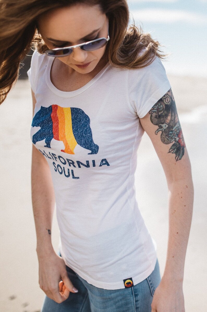 May include: A white t-shirt with a blue bear graphic and the text "CALIFORNIA SOUL". The bear has a rainbow stripe across its chest. The shirt has a scoop neck and short sleeves.