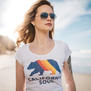 May include: A woman wearing sunglasses and a white t-shirt with a blue bear graphic and the text "CALIFORNIA SOUL".