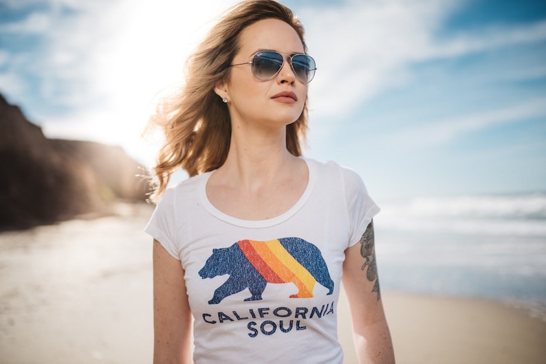 May include: A woman wearing sunglasses and a white t-shirt with a blue bear graphic and the text "CALIFORNIA SOUL".