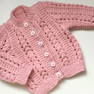 May include: A pink knitted baby cardigan with a cable knit pattern and pink flower buttons.
