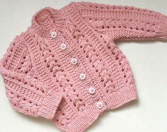 May include: A pink knitted baby cardigan with a cable knit pattern and pink flower buttons.