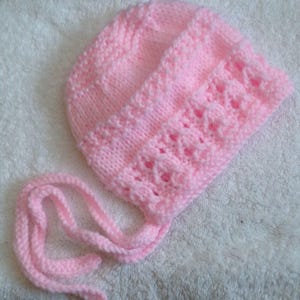 May include: A pink knitted baby hat with a tied bow at the back. The hat has a delicate lace pattern.