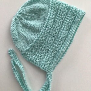 May include: A light blue knitted baby bonnet with a tie. The bonnet has a delicate stitch pattern and is made of soft yarn.