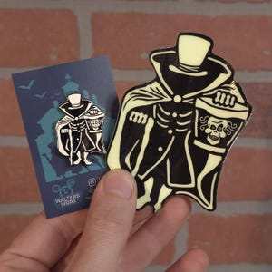 Può includere: Un adesivo e uno spillo smaltato che brillano al buio con un'illustrazione in bianco e nero di un fantasma che tiene un secchio. Il fantasma indossa un cappello a cilindro e un mantello. L'adesivo e lo spillo sono su uno sfondo blu con il testo "Walters Work" in bianco.