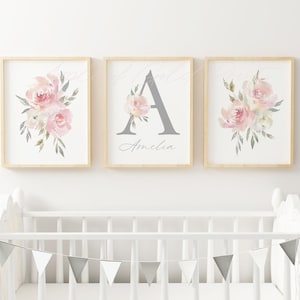 Three wall art prints in wooden frames hanging on the wall behind a white baby crib. The two outside frames are blush pink flower bouquets. The centre frame is personalised with a large grey letter and the name below it in grey cursive text.