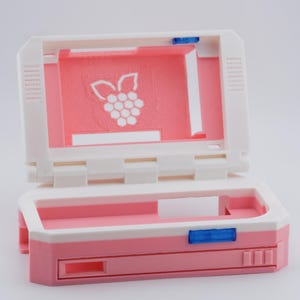 May include: A pink and white handheld gaming console with a hinged top. The screen area is pink with a white raspberry logo. The console has a rectangular shape with a slot and blue accents.