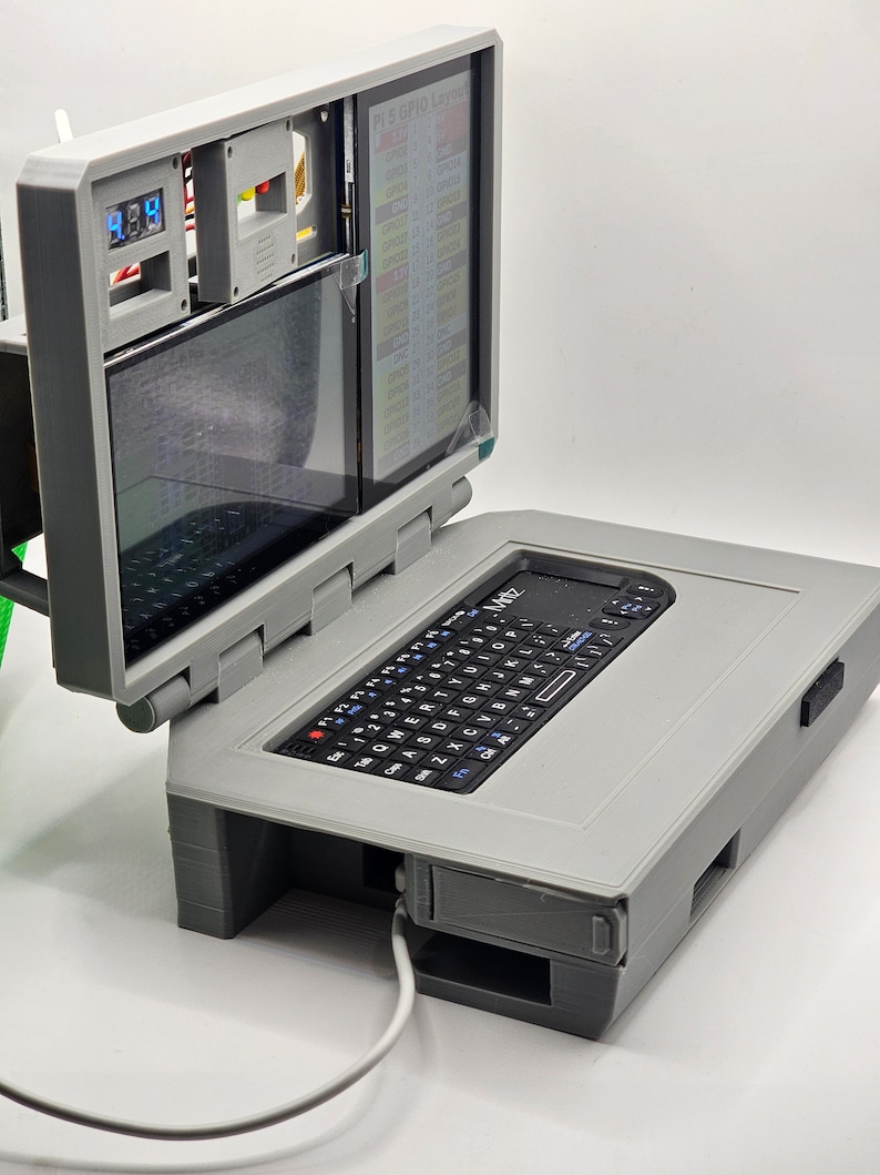 May include: A gray 3D printed laptop with a small keyboard and a screen that is open to reveal a smaller screen with text that reads "PL5 GPIO Layout".