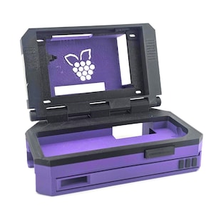 May include: A black and purple plastic computer case with a hinged lid. The interior features a cutout of a raspberry logo on the screen frame. The base includes rectangular ports and three small square buttons on the front edge of the chassis.
