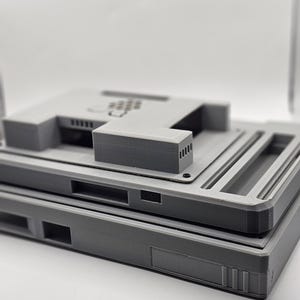 May include: Two gray 3D printed plastic cases stacked on top of each other. The top case has a rectangular opening and a series of small holes on the top.