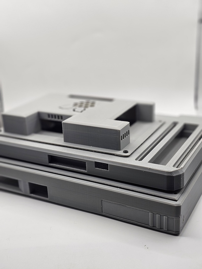 May include: Two gray 3D printed plastic cases stacked on top of each other. The top case has a rectangular opening and a series of small holes on the top.