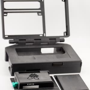 May include: A black plastic case for a Raspberry Pi computer. The case has a hinged lid and a cutout for a screen. The case is disassembled, showing the different parts.