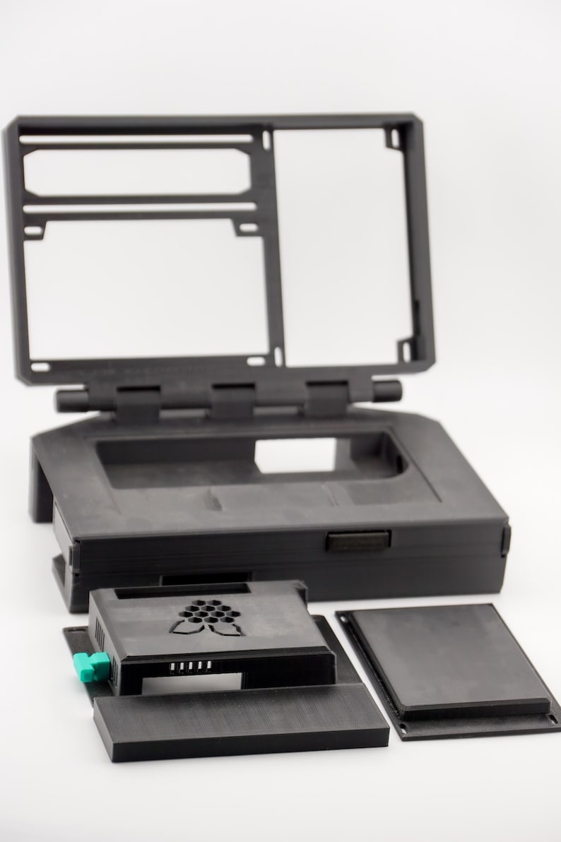 May include: A black plastic case for a Raspberry Pi computer. The case has a hinged lid and a cutout for a screen. The case is disassembled, showing the different parts.
