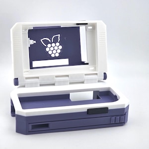 Könnte beinhalten: Ein zweifarbiges Gehäuse, weiß und lila, für elektronische Geräte. Das Gehäuse ist geöffnet und zeigt einen lilafarbenen Innenraum mit einem weißen Himbeerlogo. Das Gehäuse ist mit einem Klappdeckel und einer rechteckigen Form versehen.