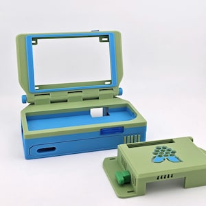 May include: A light green and blue plastic computer-style case with a hinged screen frame and a separate base cover featuring a blue honeycomb cutout design. The main unit has a rectangular opening for a screen and a hollow interior for components.