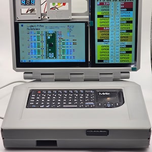 May include: A gray laptop with a black keyboard and a small touchpad labeled "Mirttz". The laptop has a screen that is open and shows a Raspberry Pi Pico Pin Reference diagram and a Pi 5 GPIO Layout diagram. The screen also shows a digital display with the number 8.8.