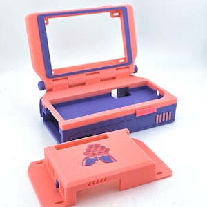 May include: A coral and purple plastic case for a small computer, featuring a hinged lid with a rectangular frame and a base with internal compartments. A separate coral cover with a purple raspberry-shaped logo is positioned in the foreground.