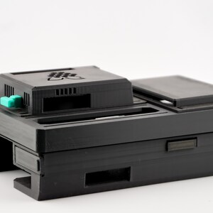 May include: A black plastic case with a green button and a slot for a device. The case has a textured surface and is designed to protect a small electronic device.