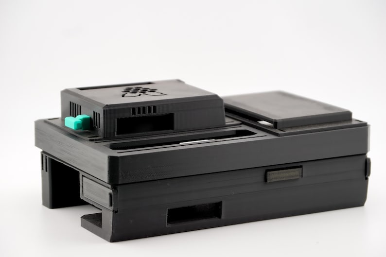 May include: A black plastic case with a green button and a slot for a device. The case has a textured surface and is designed to protect a small electronic device.