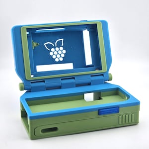 Puede incluir: Una carcasa de consola de juegos portátil de plástico azul y verde. La carcasa está abierta, revelando un interior con un logotipo de frambuesa. La carcasa tiene una tapa con bisagras y una base rectangular con un botón y ventilaciones.