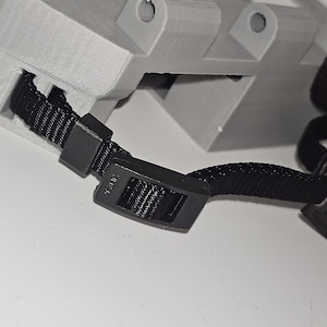 Pu&ograve; includere: Primo piano di un cinturino nero regolabile con una fibbia di plastica. Il cinturino &egrave; attaccato a un oggetto grigio stampato in 3D.