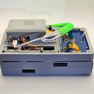 May include: A gray 3D printed case with a small fan on top, a blue circuit board with a small screen, and a green cable wrapped around the case. The case is sitting on a white surface.