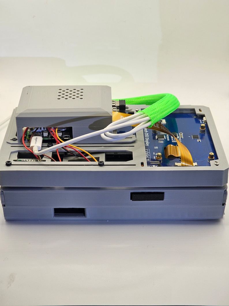 May include: A gray 3D printed case with a small fan on top, a blue circuit board with a small screen, and a green cable wrapped around the case. The case is sitting on a white surface.