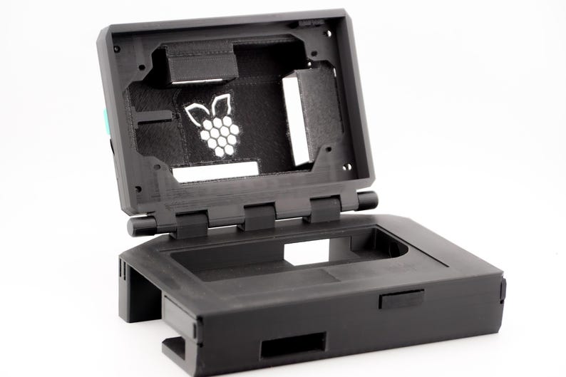 May include: A black plastic case with a hinged lid, designed to hold a Raspberry Pi computer. The lid is open, revealing the interior of the case, which has a cutout for the Raspberry Pi's board and a logo of a raspberry with leaves.