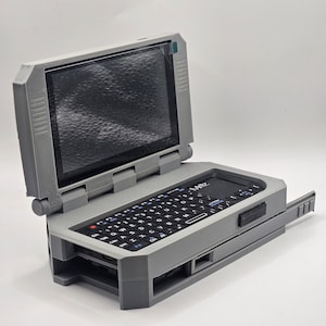 Pu&ograve; includere: Un computer portatile grigio e nero con un piccolo schermo e una tastiera completa. Il computer &egrave; aperto e lo schermo &egrave; visibile. Il computer ha un piccolo pulsante rettangolare nero sul lato destro della tastiera.