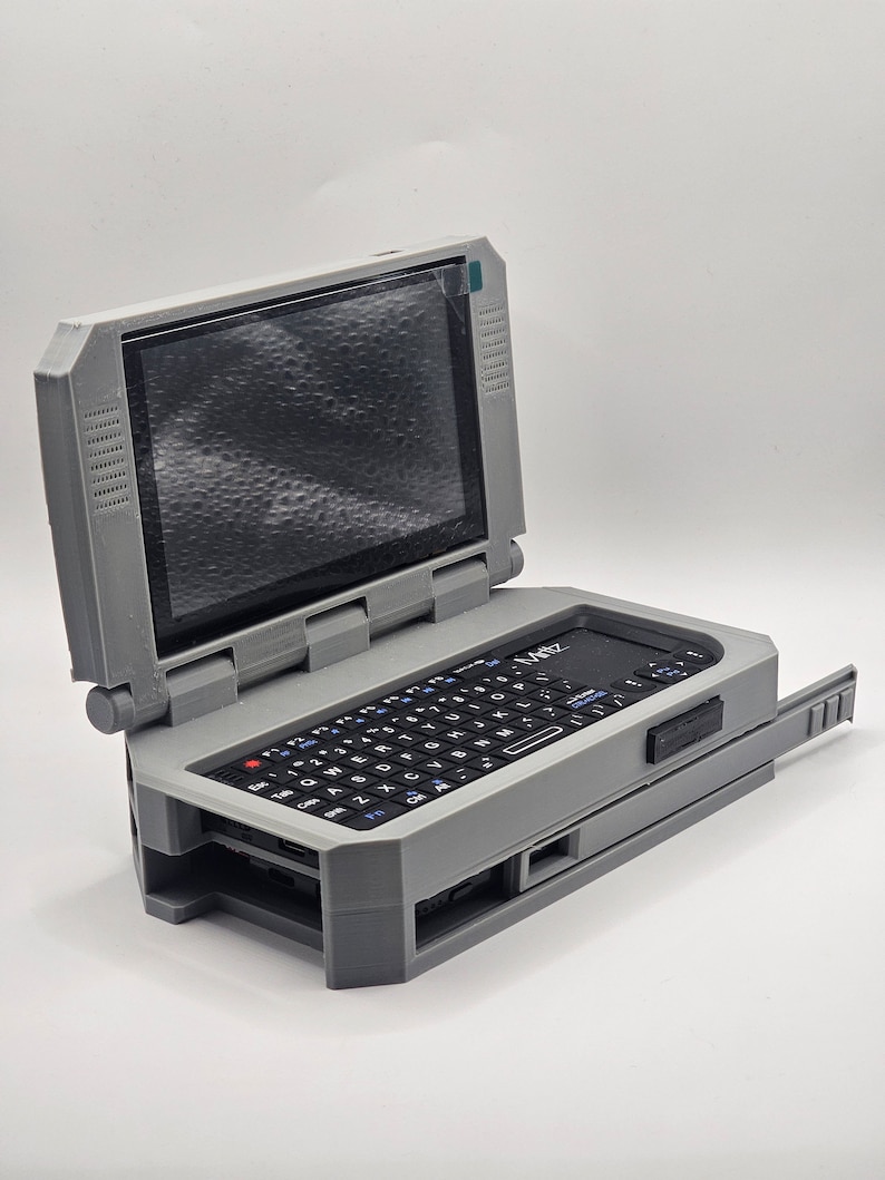 Pu&ograve; includere: Un computer portatile grigio e nero con un piccolo schermo e una tastiera completa. Il computer &egrave; aperto e lo schermo &egrave; visibile. Il computer ha un piccolo pulsante rettangolare nero sul lato destro della tastiera.