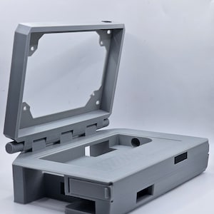 Puede incluir: Una caja gris impresa en 3D con una tapa con bisagras, diseñada para contener un pequeño dispositivo electrónico. La caja tiene varias aberturas para puertos y botones.