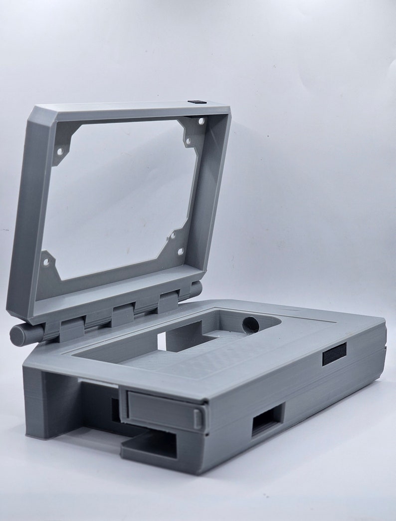 May include: A gray 3D printed case with a hinged lid, designed to hold a small electronic device. The case has several openings for ports and buttons.