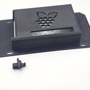 Könnte beinhalten: Ein schwarzes, 3D-gedrucktes rechteckiges Gehäuse mit einem Raspberry Pi-Logo-Ausschnitt auf der Oberseite und einer rechteckigen Öffnung an der Vorderseite. Ein kleines, separates schwarzes Kunststoffteil mit T-Profil liegt davor.