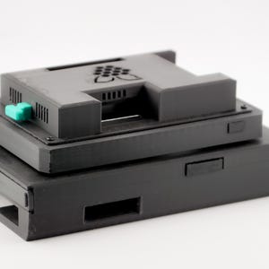 May include: A black Raspberry Pi case with a green button and a black Raspberry Pi logo. The case is stacked on top of another black case.