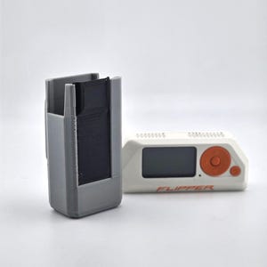 May include: A gray 3D-printed case and a white and orange electronic device. The case is rectangular with an open top and a black interior. The electronic device has a screen and the word "FLIPPER" printed on it.