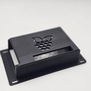 May include: A black rectangular case with a cutout design of a raspberry and honeycomb pattern. The case has a rectangular opening and mounting holes. The product is likely for electronics or a similar tech device.