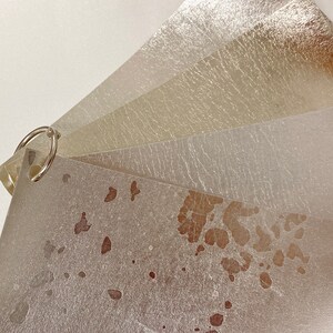 May include: A collection of metallic paper swatches in shades of silver, gold, and champagne, bound together with a silver ring. The papers have a textured, slightly distressed appearance, with some showing brown spots.