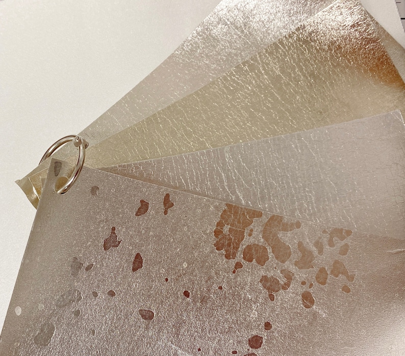 May include: A collection of metallic paper swatches in shades of silver, gold, and champagne, bound together with a silver ring. The papers have a textured, slightly distressed appearance, with some showing brown spots.